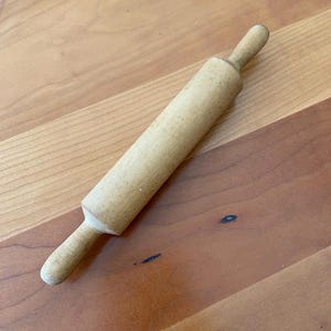 May include: A wooden rolling pin with a smooth, light brown finish. The rolling pin is approximately 12 inches long and has rounded ends.