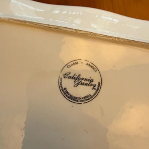 May include: A rectangular, off-white ceramic serving dish with a slightly wavy rim. The dish has a glossy finish and a circular stamp that reads "California Pantry" and "Made in China".