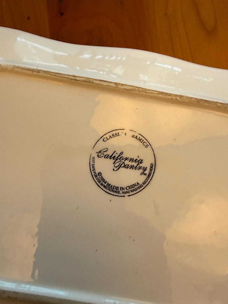May include: A rectangular, off-white ceramic serving dish with a slightly wavy rim. The dish has a glossy finish and a circular stamp that reads "California Pantry" and "Made in China".