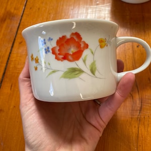 Puede incluir: Taza de cerámica blanca con un diseño floral que presenta una amapola roja grande, flores azules y amarillas más pequeñas y hojas verdes. La taza tiene un asa redondeada.
