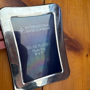 Puede incluir: Marco de fotos plateado de International Silver Company. El marco rectangular tiene esquinas redondeadas y un acabado plateado brillante. El tamaño de la foto es de 12,7 cm x 17,8 cm. El marco está sobre una superficie de madera.