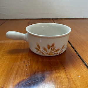 Puede incluir: Un pequeño tazón de cerámica blanco con un asa marrón y un diseño de trigo. El tazón está sobre una superficie de madera.