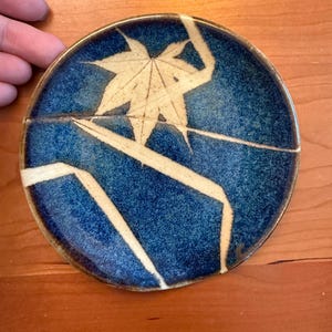 Pode incluir: Um prato redondo de cerâmica com um esmalte azul profundo e salpicado, um design proeminente de folha de bordo em cor creme e linhas angulares que se estendem pela superfície. O prato tem uma borda castanha escura e um acabamento liso e brilhante.