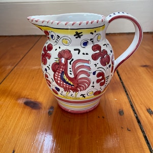 May include: A white ceramic pitcher with a red, yellow, and blue rooster design. The pitcher has a curved handle and a spout. Floral and geometric patterns decorate the body. The pitcher is approximately 7 inches tall.