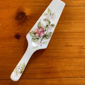 Może przedstawiać: Biały ceramiczny serwer do ciasta z kwiatowym wzorem. Serwer ma uchwyt i szerokie, płaskie ostrze. Ostrze jest ozdobione różowymi jagodami, zielonymi liśćmi i białymi kwiatami. Uchwyt ma również białe kwiaty.