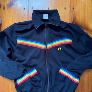 以下が含まれることがあります： 黒色のジップアップトラックジャケット。胸と袖口に虹色のストライプが入っています。ジャケットは襟付きで、フロントポケットが2つあります。小さなロゴが胸の右上に表示されています。