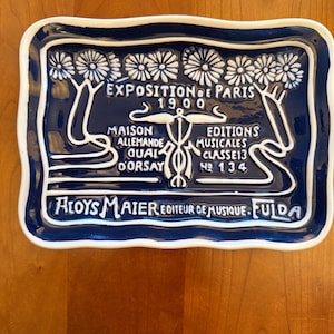 May include: A rectangular, dark blue ceramic plate with a white border and white lettering. The plate features text in French, including "Exposition de Paris 1900" and "Aloys Maier Editeur de Musique. Fulda."