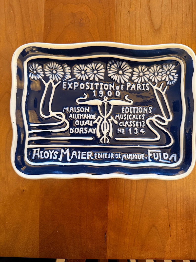 May include: A rectangular, dark blue ceramic plate with a white border and white lettering. The plate features text in French, including "Exposition de Paris 1900" and "Aloys Maier Editeur de Musique. Fulda."