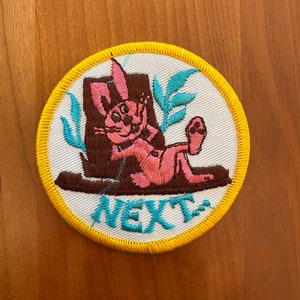 May include: A round, white embroidered patch with a yellow border. The patch features a pink rabbit sitting in a brown boot with green leaves behind it. The text "NEXT" is embroidered in blue below the rabbit.