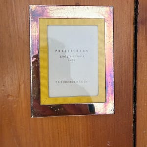 May include: A rectangular photo frame with a metallic silver border and a yellow grosgrain interior. The frame holds a white space for a 2 x 3 inch photo, with text that reads "POTTERY BARN grosgrain frame cadre" and "2 X 3 INCHES/5 X 7.6 CM".