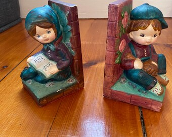 May include: A pair of bookends featuring a girl and a boy. The girl is wearing a blue dress and a blue hat and is holding a book. The boy is wearing a blue shirt and a blue hat and is holding a baseball bat. The bookends are painted in a whimsical style with a brick wall and flowers in the background.