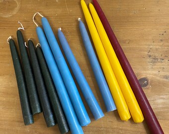 May include: A selection of taper candles in a range of colours, including dark green, blue, yellow, and burgundy. The candles are displayed on a wooden surface, highlighting their different shades and textures. The candles are of varying lengths.