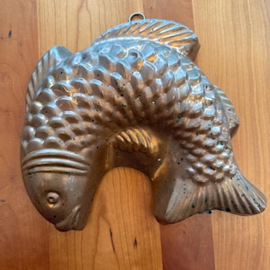 May include: A vintage copper fish-shaped mold with detailed scales and fins. The mold has a loop for hanging and is displayed on a wooden surface. The copper has a tarnished patina, adding to its antique charm.