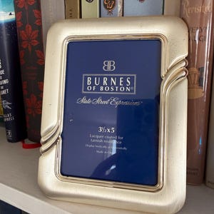 May include: A rectangular, silver-colored picture frame with rounded corners and decorative side accents. The frame has a dark blue insert with the text "BURNES OF BOSTON" and "State Street Expressions". The frame is 3.5 x 5 inches.
