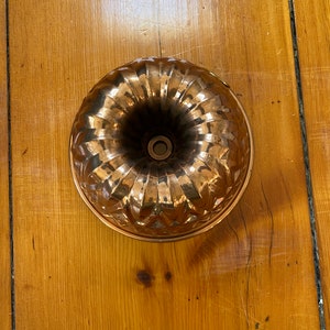 May include: A copper bundt pan with a fluted design. The pan has a central tube and a rounded base. The copper has a warm, reflective sheen, and the pan sits on a wooden surface.