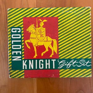 May include: A vintage box for a Golden Knight Gift Set. The box has a red, green, and yellow striped design. A knight on a horse with a shield that says "G.K." is in the center of the box.
