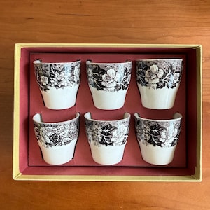 May include: A set of six white and black floral patterned egg cups in a yellow and red box. The egg cups have a curved handle and a black floral design on the top half. The box is rectangular and has a red interior.