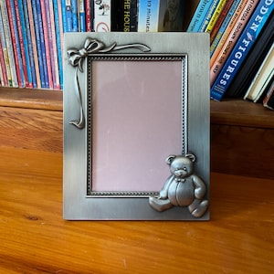 May include: Silver-toned metal picture frame with a bow and ribbon detail at the top left corner. A small teddy bear with a bow tie is on the bottom right. The frame has a pink interior and is sitting on a wooden surface.