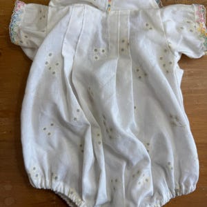 May include: White baby romper with short sleeves and a rounded collar. The romper features delicate floral embroidery and a scalloped trim with pastel colors. The fabric appears to be lightweight, suitable for warm weather.