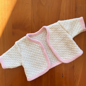 Könnte beinhalten: Eine cremefarbene Baby-Strickjacke mit offenem Vorderteil und strukturiertem Strickmuster. Die Kanten und Manschetten sind mit einer zarten rosa Bordüre versehen. Das Kleidungsstück ist für Säuglinge und Kleinkinder konzipiert.