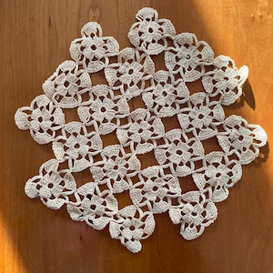May include: A delicate, off-white crocheted doily with a square shape and a floral pattern. The doily is made of fine thread and has a delicate, lacy appearance.