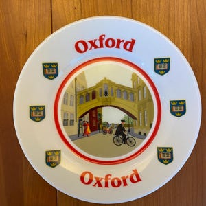 Puede incluir: Un plato de cerámica blanco con un borde rojo y blanco. El plato presenta una ilustración colorida de un puente en Oxford, Inglaterra, con el texto "Oxford" impreso en rojo en la parte superior e inferior del plato. Hay seis pequeños escudos alrededor del borde del plato.
