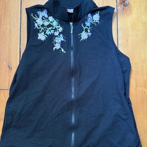 May include: A black sleeveless zip-up vest featuring a high collar and a full-length front zipper. The upper chest area is decorated with embroidered floral patterns and butterflies in shades of purple, blue, and green on both sides.