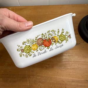 Może przedstawiać: Białe prostokątne naczynie do zapiekania z kolorowym wzorem warzywnym. Wzór zawiera pomidory, karczochy i grzyby z napisem "La Persil La Sauge". Naczynie jest trzymane w dłoni i leży na drewnianej powierzchni.