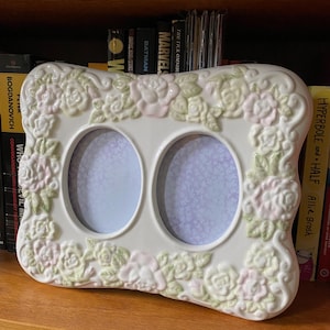 Può includere: Una cornice portafoto doppia in ceramica bianca con aperture ovali. La cornice è decorata con rose rosa e verdi in rilievo. La cornice è su una mensola di legno davanti a una libreria.