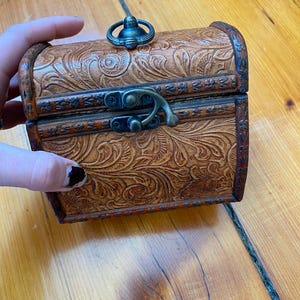 Op de afbeelding: Een kleine, houten schatkist met een bruine, lederachtige afwerking en ingewikkelde bloemmotieven. Het heeft een metalen sluiting en een decoratief handvat aan de bovenkant.