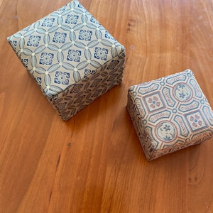 Könnte beinhalten: Zwei kleine quadratische Boxen mit aufwendigen gemusterten Stoffbezügen. Die Box auf der linken Seite hat ein hellgrünes und blaues Muster, während die Box auf der rechten Seite ein braunes, rotes und blaues Muster hat.