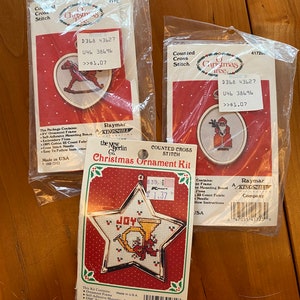 Op de afbeelding: Drie geteld kruissteek kerstversiering sets. De sets bevatten een schommelpaard, de kerstman en een ster met een trompet. Elke set bevat een frame, montageplaat, garen, stof, naald en instructies. De sets zijn verpakt in doorzichtige plastic zakken met prijskaartjes.