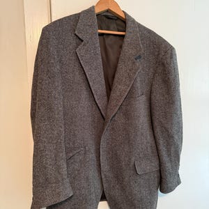 May include: A gray herringbone blazer hanging on a wooden hanger. The blazer has a notched lapel, two front pockets, and a single-breasted closure. The fabric has a subtle pattern and a classic, tailored look.