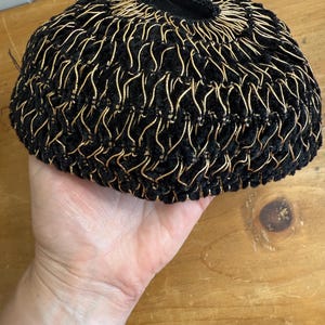以下が含まれることがあります： 黒と金の織り帽子。丸みを帯びた形で、中央に開口部があります。黒い生地に金色の金属糸が織り込まれたテクスチャードデザインが特徴です。帽子は手で持たれています。