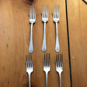 Puede incluir: Seis tenedores plateados dispuestos sobre una superficie de madera. Los tenedores tienen un diseño clásico con púas y mangos decorativos. La disposición resalta la elegante forma de la cubertería, adecuada para comer.