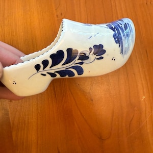 May include: A white ceramic Dutch clog with blue floral and abstract designs. The decorative shoe features a curved shape and is painted with dark blue accents. The clog is a traditional souvenir or decorative item.