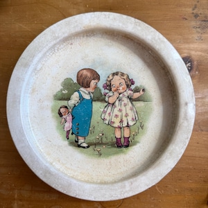 Op de afbeelding: Vintage ronde serveerschaal met een geschilderde illustratie van drie kinderen in een tuin. De schaal heeft een crèmekleurige rand en een licht verweerde uitstraling, wat bijdraagt aan de antieke charme. De illustratie toont kinderen in vintage kleding.