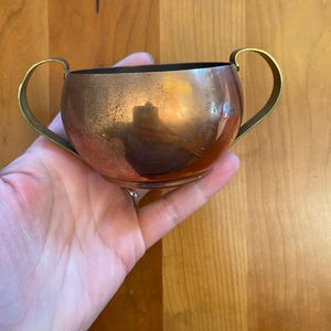 May include: A small, copper-colored bowl with two brass handles. The bowl has a rounded shape and a dark interior. The copper has a reflective surface, and the brass handles are curved. The bowl is held in a hand.