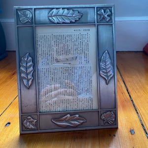 May include: A rectangular metal picture frame with a leaf design. The frame has a silver finish and features embossed leaf details on each corner and side. The frame holds a page of Japanese text.