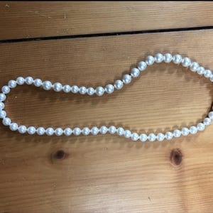 May include: A strand of white faux pearls, displayed on a wooden surface. The pearls are round and evenly spaced, creating a classic necklace design. The necklace is a simple, elegant accessory.