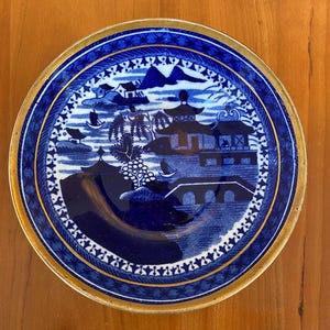 May include: A blue and gold ceramic plate with a Chinese-inspired design. The plate features a pagoda, a bridge, and a mountain range. The border of the plate is decorated with a gold trim.