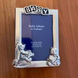 May include: Silver-toned baby photo frame with a blue background. The frame has a "BABY" inscription and features two teddy bears, one riding a rocking horse and the other holding a book. The frame is designed to hold a 2x3 inch photo.