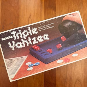 May include: Deluxe Triple Yahtzee game box with dice and game pieces on a wooden surface. The box features the game title and the tagline "Three times the strategy, fun and excitement for Yahtzee enthusiasts."
