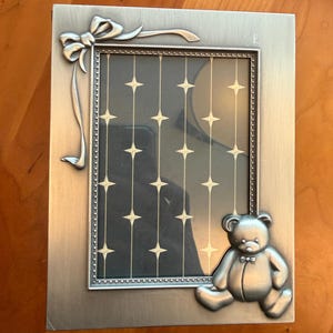 May include: A rectangular silver-toned picture frame with a bow and ribbon detail at the top left corner and a teddy bear at the bottom right. The frame has a dark background with a star pattern.