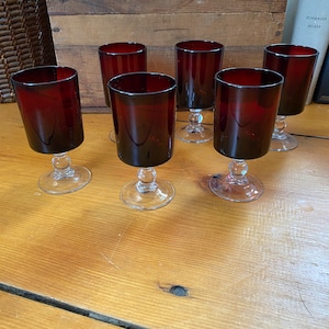 Puede incluir: Seis vasos de vidrio transparente con tallo y base de color rojo oscuro. Los vasos están dispuestos en fila sobre una superficie de madera.