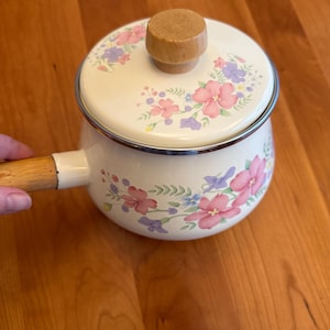 May include: Cream-colored saucepan with a lid, featuring a floral pattern in pink, purple, and blue. The handle and lid knob are made of wood. The pot has a silver rim and is designed for cooking.