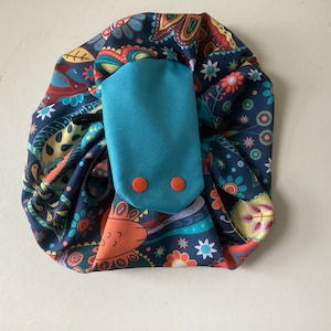 May include: A circular drawstring bag with a colourful paisley design in navy, teal, orange, and yellow. It has a teal flap secured by two orange buttons. The bag is for storage.