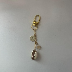 May include: Gold-toned keychain with a lobster clasp. The keychain features a chain with a circular charm with the letter "N", a small flower charm, and a white and gold seashell charm. The background is a solid white.