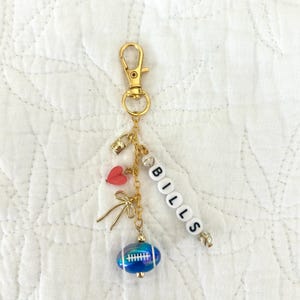May include: A gold-colored keychain with a lobster clasp, featuring charms like a small lock, a red heart, and a bow. The word "BILLS" is spelled out in white letter beads. A blue and white football charm hangs at the bottom.