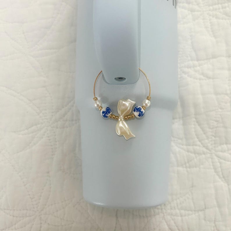 May include: A gold-colored wine glass charm with a cream-colored bow-shaped pendant and blue and white ceramic beads. The charm is attached to a light blue tumbler.
